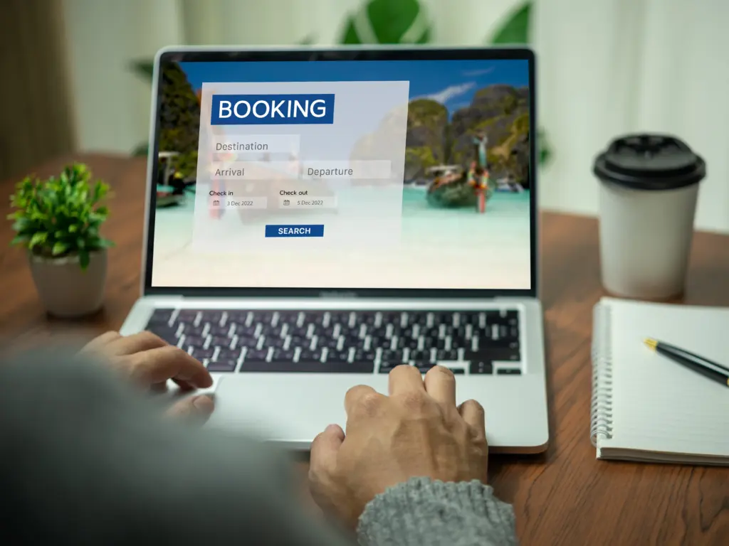 Visualisierung Lokale SEO. Person nutzt einen Laptop mit geöffneter Buchungsseite für Reisen; im Hintergrund der Website ist ein tropischer Strand mit Booten zu sehen. Auf dem Tisch stehen eine Pflanze, ein Coffee-to-go-Becher und ein Notizbuch mit Stift.